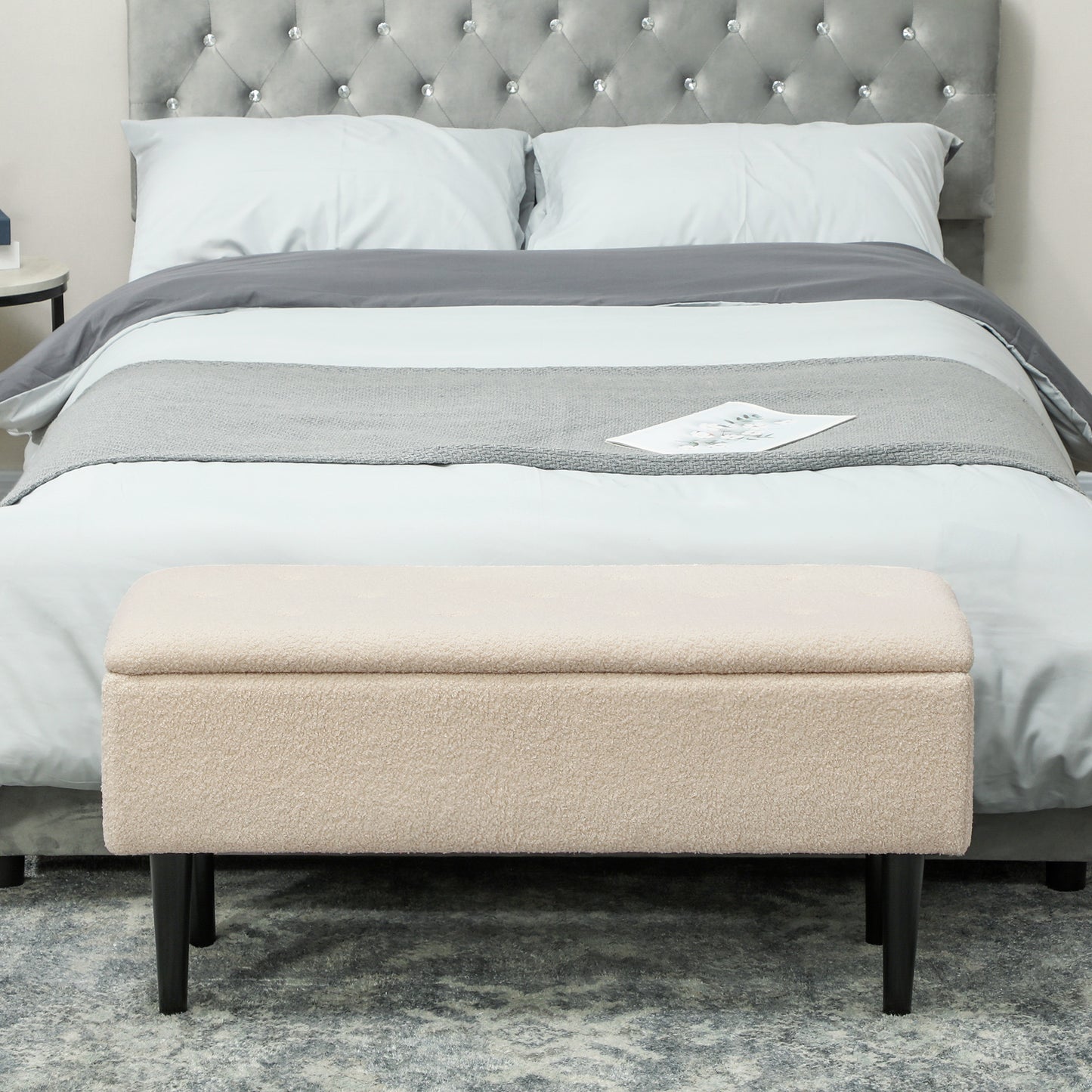 Cream teddy fleece storage ottoman with black legs placed at the foot of a bed