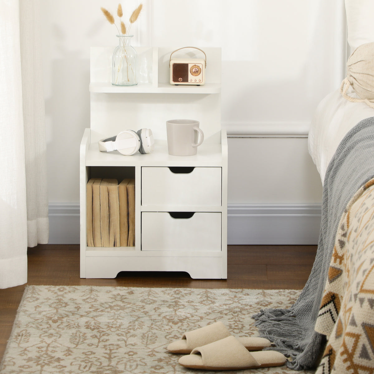 White bedside table beside a bed, with two drawers, an open cubby, and upper shelf