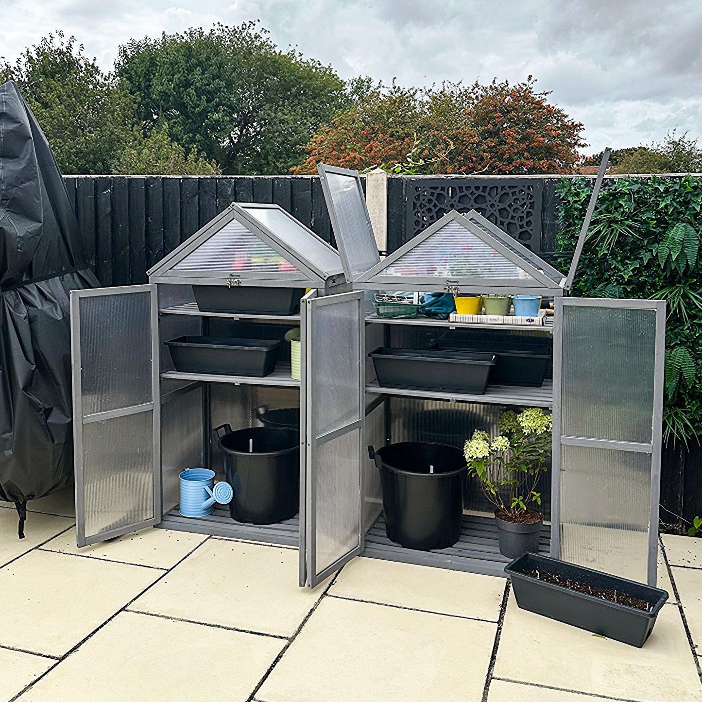 Grey greenhouse with open roof panels and double doors, holding plants and gardening supplies
