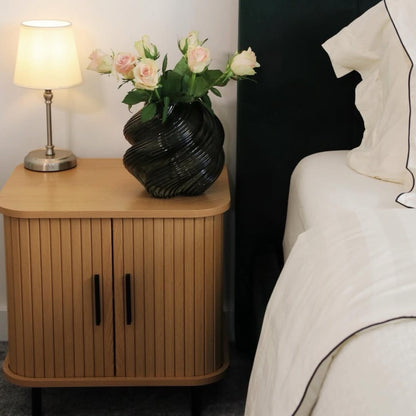 Oak-tone nightstand with vertical slatted doors and black handles beside a bed
